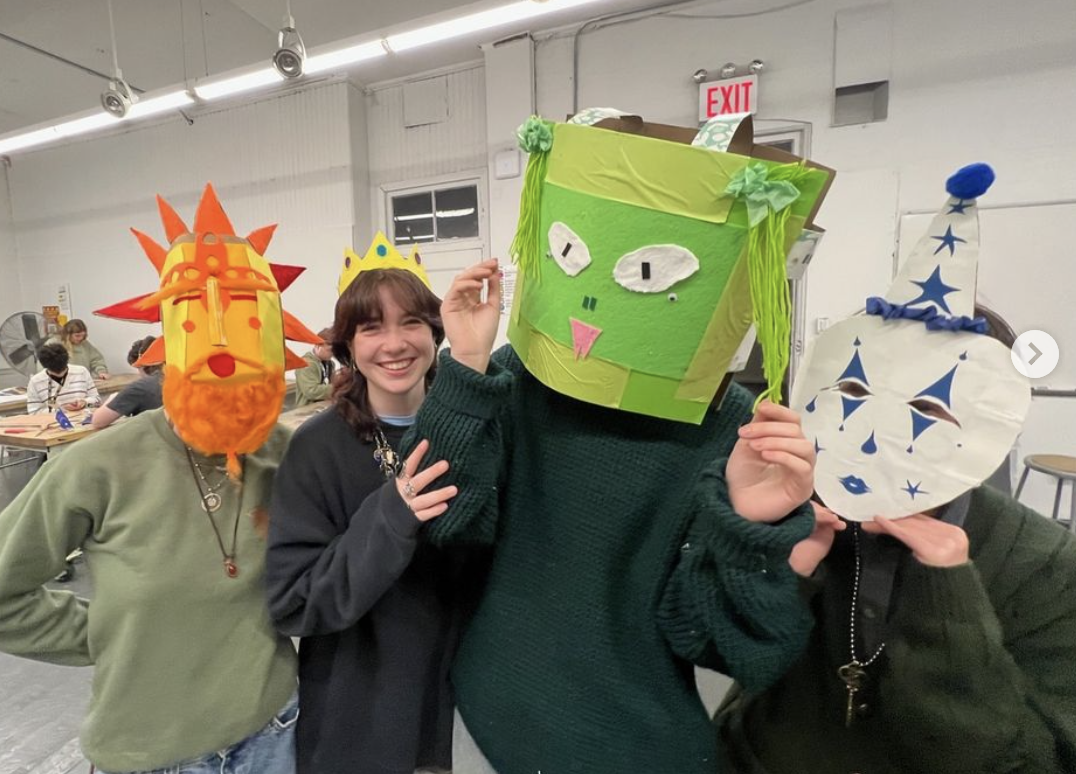 Four individuals wearing handmade masks. One mask resembles a bright orange sun with pointed rays, another is a large green box-like face with dangling strands, and a third features a white and blue clown-inspired design. The fourth person, in the middle, smiles without a mask and wears a yellow crown. Tables and other participants are visible in the background.